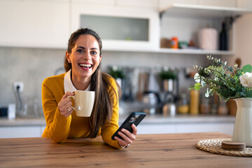 Portrait of happy young woman using mobile phone and having coffee while looking at camera in the...
