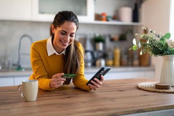 Charming young woman looking at credit card using mobile phone secured online e-banking app,...