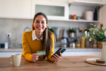Smiling businesswoman looking at camera holding credit card and mobile phone at home.