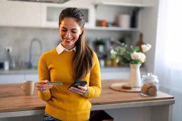 Cheerful female customer with mobile phone looking at credit card while standing at home.