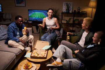 Wide shot of four diverse young friends relaxing in low-beamed living room talking and enjoying...