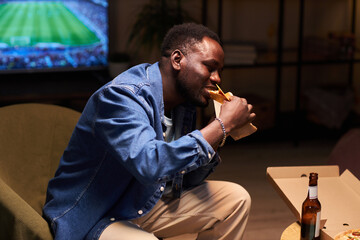 Medium full shot of young Black man biting delicious taco while sitting on armchair at home