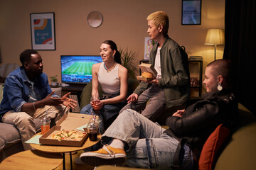 Wide shot of four diverse young adults sitting in living room with fast food and soft drinks while...