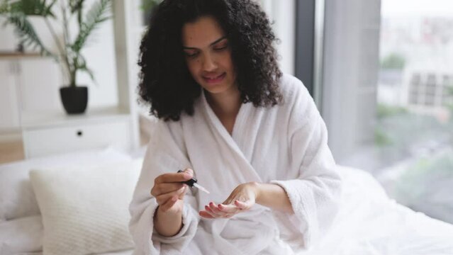 Close up view of hands of multinational young lady in dressing gown, paints nails while sitting on soft bed in bright room. Joyful housewife increasing life satisfaction by making manicure with polish