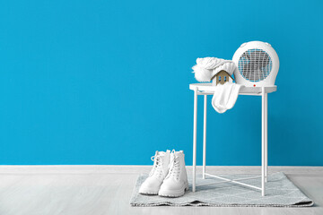 Electric fan heater with hat, socks on coffee table and boots near blue wall. Concept of heating...