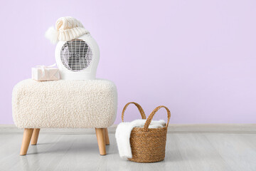 Electric fan heater with hat, gift box on pouf and basket near lilac wall. Concept of heating season