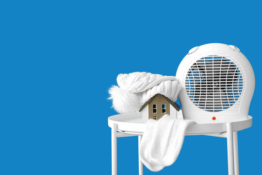 Electric fan heater with decorative house, hat and socks on coffee table against blue background. Concept of heating season
