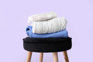 Stacked sweaters with gloves on stool against lilac background