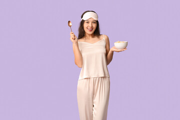 Beautiful young happy Asian woman in pajamas with sleeping mask and bowl of cornflakes on purple...
