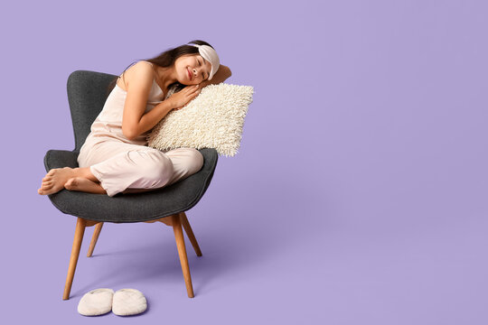 Beautiful young Asian woman in pajamas with sleeping mask and pillow sleeping on armchair against purple background - Powered by Adobe