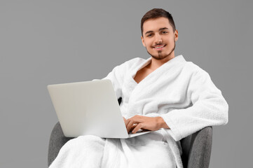 Handsome young man in bathrobe with laptop on grey background