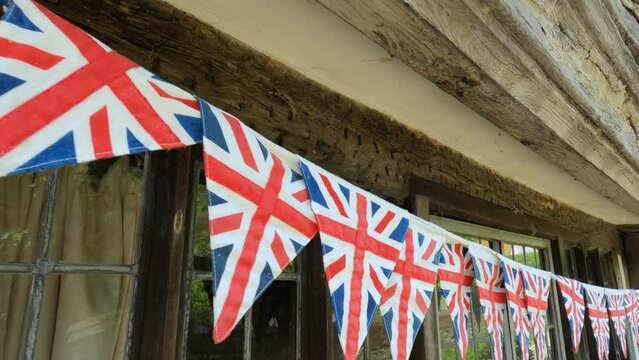 CLOSE UP: String of Union Jack flags strewn under wooden beams of an old house. Bunting English pennant as a symbol of a festive atmosphere and a patriotic pride in historic town during celebration.