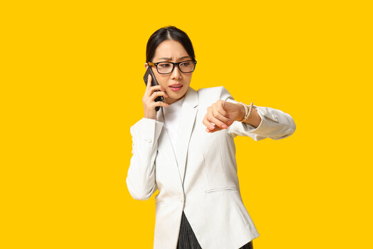 Portrait of young Asian businesswoman talking by phone and checking time on yellow background