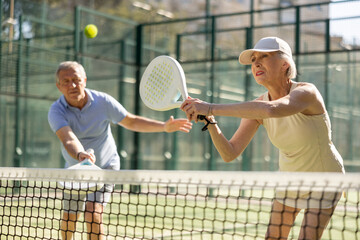 Sportive elderly man and woman engaged in Padel Tennis in open-air court of tennis club