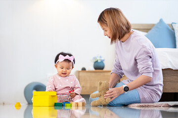 Asian mother is playing with her pretty smiling baby daughter with wooden toy block while spending quality time in the bed for family happiness and parenting