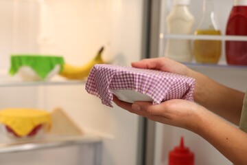Woman holding bowl covered with beeswax food wrap near refrigerator in kitchen, closeup