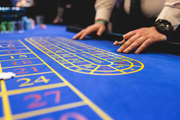 Vibrant casino table with roulette in motion, with casino chips, tokens, the hand of croupier, dollar money and a group of gambling rich people playing bet, blue poker table and deck of cards