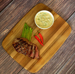 Wooden plate with a piece of fried beef tenderloin with asparagus, hot peppers and sauce.