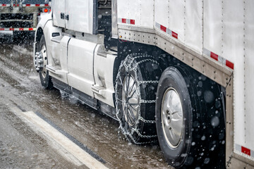 .Chains for better grip on slippery road surfaces on a white big rig semi truck drive axle wheel transporting cargo in dry van semi trailer running at winter weather during a snow storm