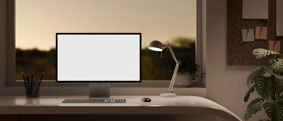 Modern home office in the evening with a white-screen computer on a desk near by the window.