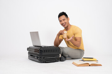 Positive Asian man is sitting on an isolated white background with his laptop, books, and a luggage.