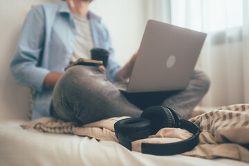 Fototapeta premium Focus on headphone. People sitting on bed holding coffee glass and using computer laptop. Freelancer working online at home. Meeting video conference with customer or connecting with social network