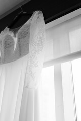 Black and white portrait of a lace wedding dress with long sleeves hanging up against the window.