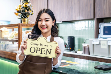 Happy beautiful Asian female waitress or younger shopkeeper in an apron holding open sign while standing at cafe or in restaurant, with pleasured smiling, welcome, business owner startup ideas - obrazy, fototapety, plakaty