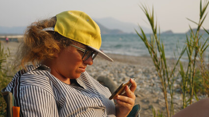 A woman wearing glasses and a yellow baseball cap is sitting on a beach watching a film on her...
