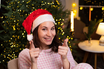 A young attractive woman in cozy pajamas is sitting at home on a chair in a red Christmas hat, smiling and pointing two fingers at the camera