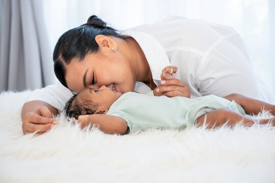 Asian mother kissing her 1-month-old baby newborn son, half Thai half Nigerian who is lying in white bed, with smile and happy, to relationship in family and infant concept.
