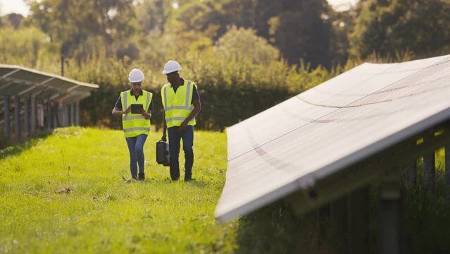 Male and female engineers wearing hard hats and hi vis safety vests with digital tablet walking through field outdoors inspecting solar panels on sustainable energy farm - shot in slow motion