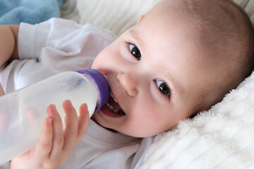 A child drinks milk from a baby bottle. Baby girl, top view, in white clothes, European appearance. Nutrition, choice of natural or artificial feeding.