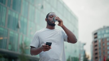 Relaxed african american man drinking coffee in paper cup enjoying walking in city downtown near modern building. Bearded guy in white t-shirt strolling outdoors. Urban lifestyle, morning, breakfast.