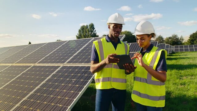 Male and female engineers wearing hard hats and hi vis safety vests with digital tablet outdoors inspecting solar panels on sustainable energy farm - shot in slow motion