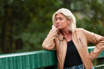 An older blonde woman leans on a bridge railing and looks into the stream below.