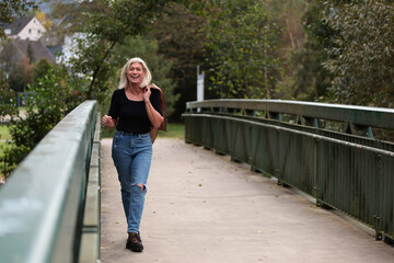 An older blonde woman in a leather jacket and a black shirt casually walks across a bridge with a...