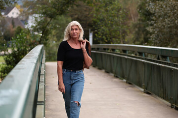 An older blonde woman in a leather jacket and a black shirt casually walks across a bridge with a...