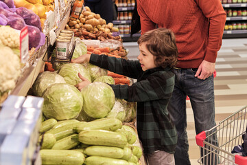 In the grocery aisle, a young child reaches for a head of lettuce under the watchful eye of an adult, engaging in the choice of fresh vegetables. This scene is a slice of daily life, showing early