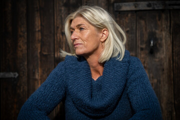 Woman blonde head portrait in her mid-fifties wearing a wide blue knitted sweater, sitting in front...