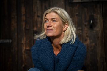 Woman blonde head portrait in her mid-fifties wearing a wide blue knitted sweater, sitting in front of a wooden door and looking curiously into the distance