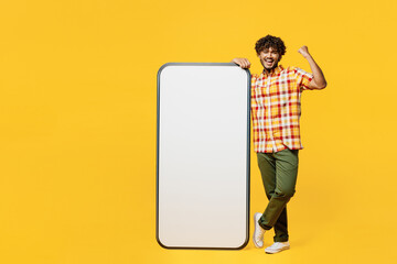 Full body young Indian man wear shirt casual clothes big huge blank screen mobile cell phone smartphone with mockup area do winner gesture isolated on plain yellow color background. Lifestyle concept
