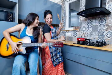 Indian mother and daughter in kitchen having fun. Girl playing Guitar and mother dancing Having...