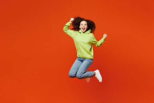 Full body young woman of African American ethnicity she wear green hoody casual clothes jump high do winner gesture celebrate clenching fist isolated on plain red orange background. Lifestyle concept