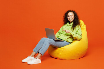 Full body smiling young IT woman of African American ethnicity in green hoody casual clothes sit in...
