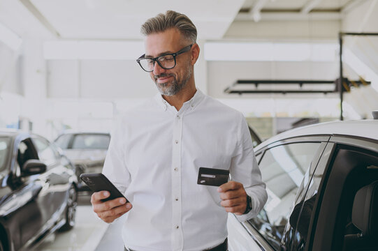 Adult man customer male buyer client wear shirt use mobile cell phone hold credit card choose auto want to buy new automobile in car showroom vehicle salon dealership store motor show. Sales concept.