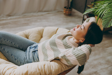 Young woman of Asian ethnicity wear casual clothes holding hands behind neck close eyes sits on armchair stay at home hotel flat rest spend free spare time in living room indoor People lounge concept - obrazy, fototapety, plakaty