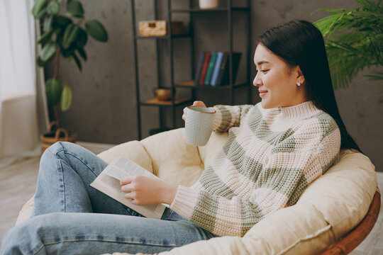 Side view young happy smart fun woman wear casual clothes sits on armchair read book novel drink tea stay at home hotel flat rest relax spend free spare time in grey living room indoor Lounge concept