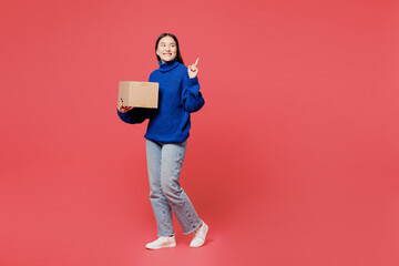 Full body young happy woman of Asian ethnicity she wearing blue sweater casual clothes hold cardboard box point aside isolated on plain pastel light pink background studio portrait. Lifestyle concept.
