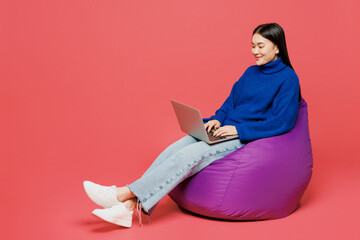 Full body young IT woman of Asian ethnicity she wear blue sweater casual clothes sit in bag chair hold use work on laptop pc computer isolated on plain pastel light pink background. Lifestyle concept
